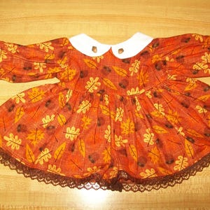 Könnte beinhalten: Ein braunes und oranges Kleid mit weißem Kragen und brauner Spitzenborte. Das Kleid hat ein Muster aus Blättern und Eicheln.