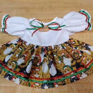 Pode incluir: Vestido branco para boneca com um laço de fita vermelho e verde e um botão de gato. A saia é estampada com um padrão de gatos usando chapéus de Papai Noel.