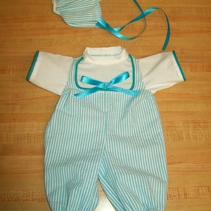 May include: A blue and white striped baby outfit with a matching hat. The outfit has a white top and blue and white striped overalls with a turquoise ribbon bow. The hat is also blue and white striped.