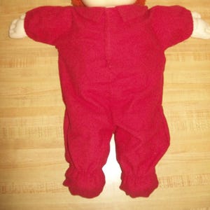 May include: A red doll wearing a red onesie with a collar. The doll has white hands and a white face. The doll has orange hair.