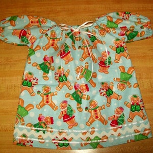 May include: A light blue dress with short sleeves, patterned with gingerbread people in festive outfits. The gingerbread men have red, green, and white details. The dress has a white ribbon and a white scalloped trim along the bottom.