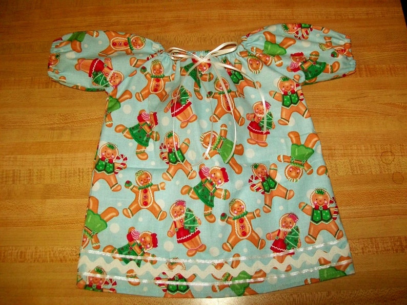 May include: A light blue dress with short sleeves, patterned with gingerbread people in festive outfits. The gingerbread men have red, green, and white details. The dress has a white ribbon and a white scalloped trim along the bottom.