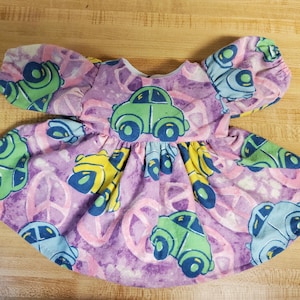 May include: Purple doll's dress with a peace sign and car print. The dress has puffed sleeves and a gathered skirt.