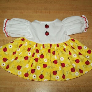 Könnte beinhalten: Ein weißes Puppenkleid mit roten Besätzen an den Ärmeln und einem gelben Rock mit Marienkäfer-Druck. Das Mieder des Kleides hat zwei rote Knöpfe.