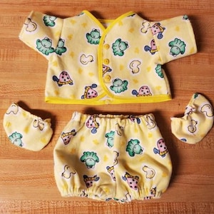 Peut inclure: Un ensemble de vêtements de bébé jaune avec un motif de grenouille et de tortue. L'ensemble comprend un haut cache-cœur à manches courtes, un bloomer et deux chaussons. La bordure est jaune et le tissu est orné de grenouilles, de tortues et d'autres formes.
