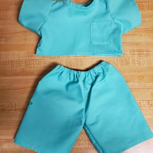 May include: A teal blue scrub top and matching trousers for a stuffed animal. The top has a small pocket on the front.