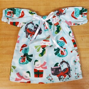 May include: A light blue Christmas dress with short sleeves and a white ribbon tie. The dress has a repeating pattern of festive cats wearing hats and scarves, with snowflakes and Christmas lights. The fabric is soft and appears to be a lightweight material.