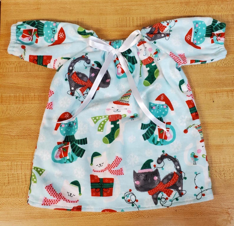 May include: A light blue Christmas dress with short sleeves and a white ribbon tie. The dress has a repeating pattern of festive cats wearing hats and scarves, with snowflakes and Christmas lights. The fabric is soft and appears to be a lightweight material.