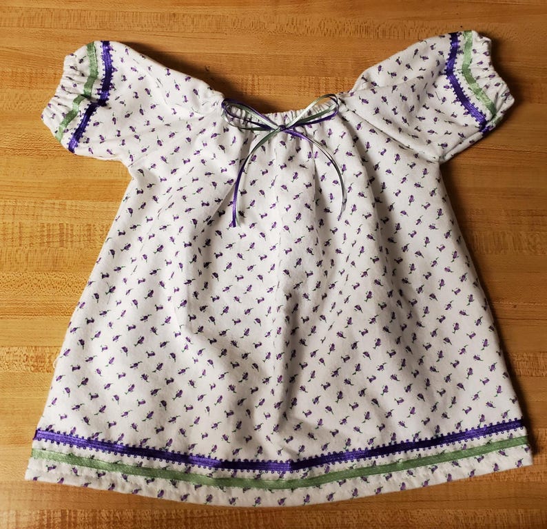 May include: White baby dress with a small purple floral pattern. The dress features short sleeves with green and purple trim, a green and purple ribbon around the hem, and a bow at the neckline. The dress is made of a lightweight fabric.