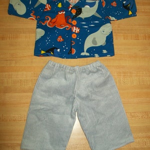 Pode incluir: Uma camisa azul e branca de manga curta com uma estampa de várias criaturas marinhas, incluindo um polvo, uma baleine e peixes. A camisa tem um fecho de botões na frente e é combinada com calças de veludo cotelê cinzas.