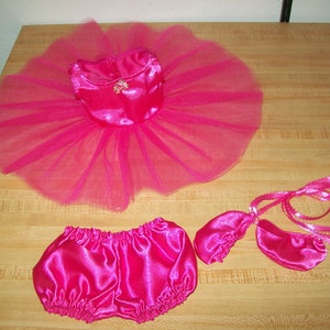 May include: A pink satin and tulle ballerina outfit for a doll. The outfit includes a dress, bloomers, and shoes. The dress has a satin bodice and a tulle skirt. The bloomers are made of satin and have a ruffled edge. The shoes are made of satin and have a ribbon tie.