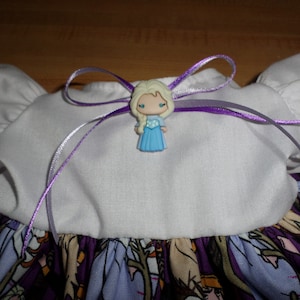 May include: A white and purple dress with a cartoon character embellishment. The dress features a white bodice with puffed sleeves and a purple ribbon bow. The skirt has a colorful print with cartoon characters.