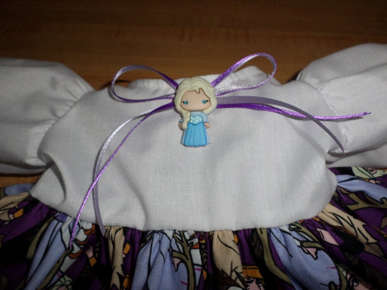 May include: A white and purple dress with a cartoon character embellishment. The dress features a white bodice with puffed sleeves and a purple ribbon bow. The skirt has a colorful print with cartoon characters.