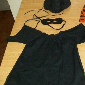 Pode incluir: Um traje de bruxa preto para crianças, incluindo um chapéu de bruxa preto, uma capa preta e uma máscara preta para os olhos.