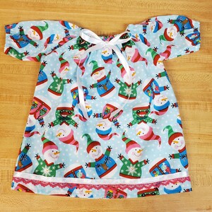 May include: A light blue dress with short sleeves, patterned with smiling snowmen in different outfits. It has a white ribbon tie and a red lace trim. The dress is made of a soft, lightweight fabric, suitable for warmer weather. The dress is approximately 60 cm in length.