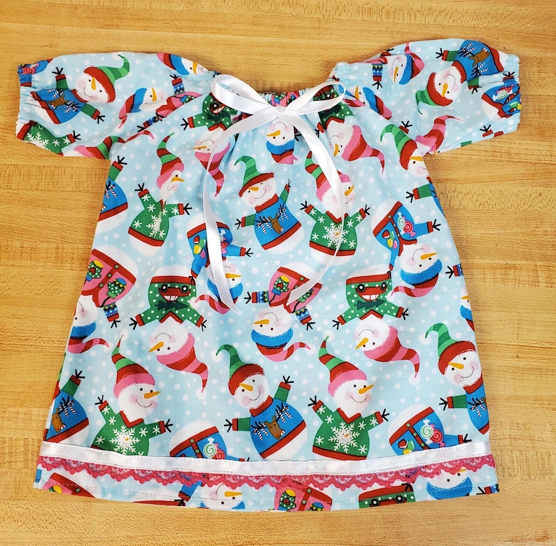 May include: A light blue dress with short sleeves, patterned with smiling snowmen in different outfits. It has a white ribbon tie and a red lace trim. The dress is made of a soft, lightweight fabric, suitable for warmer weather. The dress is approximately 60 cm in length.