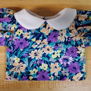 Könnte beinhalten: Eine kurzärmelige Bluse mit dunkelblauer Basis und einem lebendigen Blumenmuster in Violett-, Gelb- und Pfirsichtönen. Das Kleidungsstück verfügt über einen einfarbigen weißen Rundkragen und voluminöse Puffärmel, die eine markante Silhouette erzeugen.