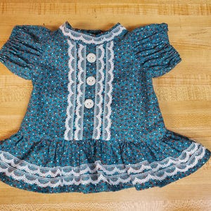 Könnte beinhalten: Ein blaugrünes Kleid mit kurzen Ärmeln und einem kleinen Blumenmuster mit weißen und roten Akzenten. Das Kleid hat einen hohen Ausschnitt, drei weiße Knöpfe in der Mitte und eine weiße, gewellte Spitzenborte am Kragen, Oberteil und Saum.