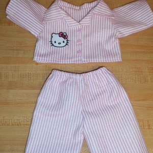 May include: A doll's pink and white striped pyjama set. The top features a collar, buttons, and a Hello Kitty motif. The shorts have an elasticated waist. The fabric is a soft cotton blend.