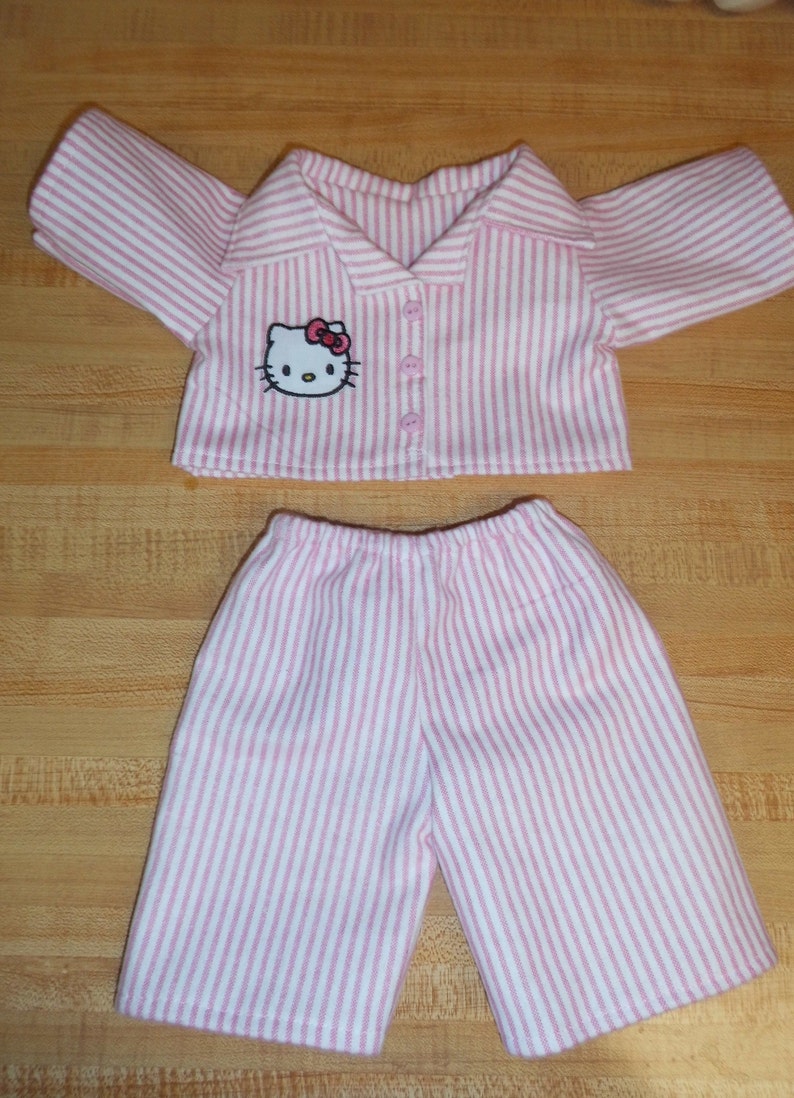 May include: A doll's pink and white striped pyjama set. The top features a collar, buttons, and a Hello Kitty motif. The shorts have an elasticated waist. The fabric is a soft cotton blend.
