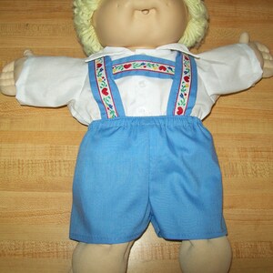 May include: A Cabbage Patch Kid doll wearing a white button-down shirt with blue and white suspenders and blue denim shorts. The suspenders have a red, white, and blue floral pattern.