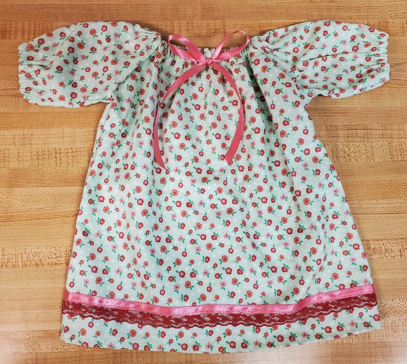 May include: A light blue floral print dress with short, puffed sleeves and a pink ribbon tie at the neckline. The dress features a pink lace trim at the hemline, creating a vintage aesthetic.
