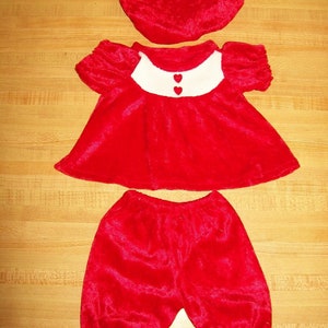 May include: A red velvet doll outfit with a hat, dress, and pants. The dress has a white collar with two red heart buttons.