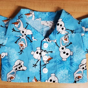 May include: A light blue button-up shirt featuring a repeating pattern of the snowman Olaf from the movie Frozen. The shirt has short sleeves and white buttons. The background is a light blue color with snowflakes.