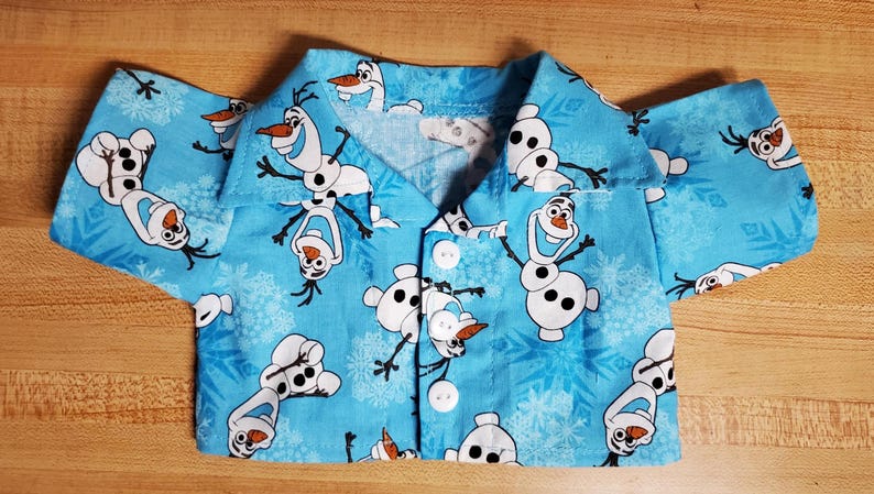 May include: A light blue button-up shirt featuring a repeating pattern of the snowman Olaf from the movie Frozen. The shirt has short sleeves and white buttons. The background is a light blue color with snowflakes.