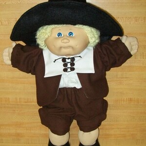Puede incluir: Una muñeca Cabbage Patch Kid vestida con un traje de peregrino marrón con un sombrero negro y un cuello blanco. La muñeca tiene el pelo rubio y los ojos azules.