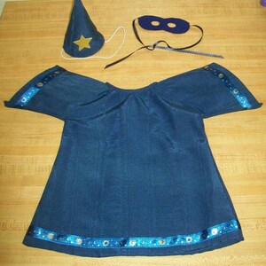 May include: A dark blue wizard costume set, including a tunic, hat, and eye mask. The tunic has blue and silver trim along the sleeves and hem. The hat is cone-shaped with a gold star. The eye mask is dark blue with black ties.