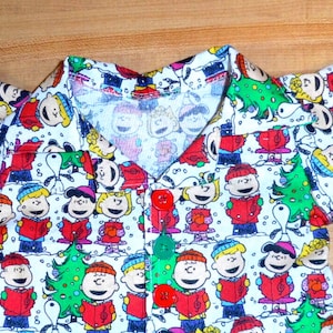 May include: A white button-down shirt with a Christmas-themed print featuring characters from the Peanuts comic strip. The shirt has red buttons and a red collar.