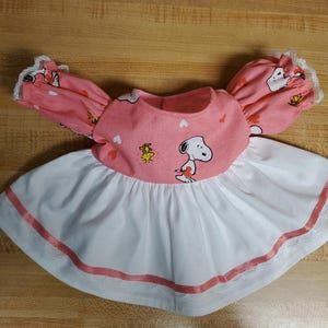 Può includere: Un vestito per bambole rosa e bianco con una stampa di Snoopy e Woodstock. Il vestito ha un bordo di pizzo bianco e un nastro rosa intorno alla parte inferiore.