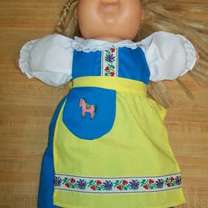 May include: A Cabbage Patch Kid doll wearing a blue and yellow dress with a white lace collar and a pink horse applique. The dress has a yellow apron with a floral border.