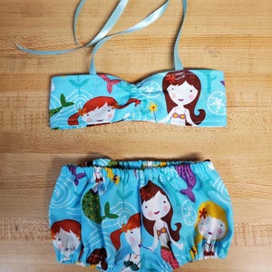May include: A two-piece baby swimsuit set featuring a light blue fabric with a mermaid pattern. The top is a bandeau style with light blue ribbon straps. The bottoms are ruffled, with an elastic waistband. The swimsuit is designed for babies and toddlers.
