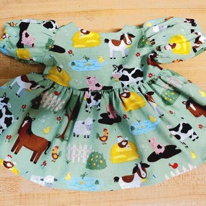 May include: A light green baby dress with a farm animal print. The dress has a gathered skirt and short, puffed sleeves. The print includes cows, horses, pigs, sheep, chickens, and hay bales.