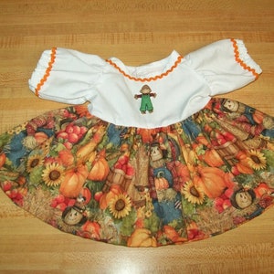 May include: A white dress with orange trim and a scarecrow applique. The skirt is made of a fabric with a fall-themed print featuring pumpkins, sunflowers, and scarecrows.