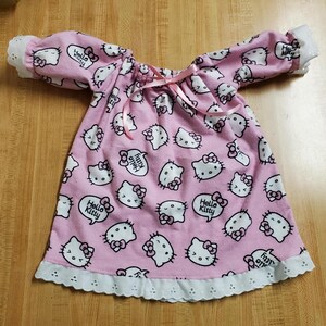 May include: A pink nightdress patterned with white Hello Kitty faces and speech bubbles saying "Hello Kitty". It has long sleeves with white lace cuffs and a white lace hem. A pink ribbon is tied at the neckline. The nightdress is made of soft fabric.
