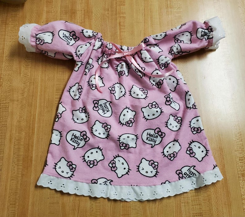 May include: A pink nightdress patterned with white Hello Kitty faces and speech bubbles saying "Hello Kitty". It has long sleeves with white lace cuffs and a white lace hem. A pink ribbon is tied at the neckline. The nightdress is made of soft fabric.
