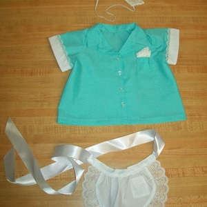 May include: A turquoise blue shirt with white trim and a white lace apron with a white ribbon. The shirt has a collar and buttons. The apron has a lace trim and a pocket.