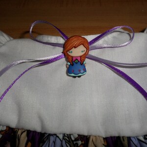 May include: A decorative bow featuring a cartoon character with red hair, a blue dress, and a purple ribbon. The bow is attached to a white fabric with a floral pattern at the bottom. The ribbon is a vibrant purple color.