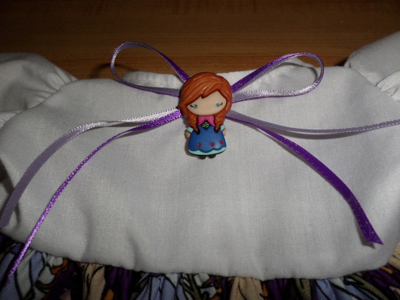 May include: A decorative bow featuring a cartoon character with red hair, a blue dress, and a purple ribbon. The bow is attached to a white fabric with a floral pattern at the bottom. The ribbon is a vibrant purple color.