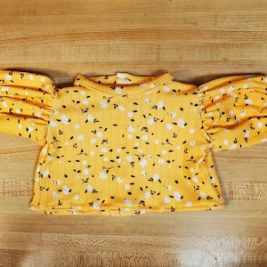 May include: A yellow crop top with a floral pattern of white flowers and black leaves. The top has a ribbed texture and short, puffed sleeves. The neckline is a simple, rounded style.