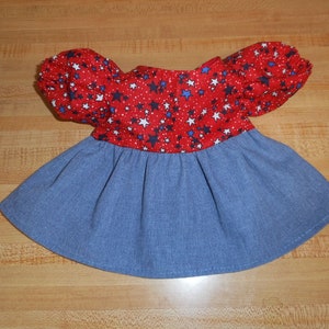 May include: A doll dress with a red bodice featuring white and blue stars on a white dotted background. The dress has puffy sleeves and a blue denim skirt. The dress is designed for a doll.