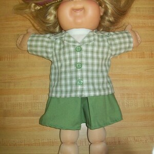 May include: A doll with blonde hair, brown eyes, and a round face. The doll wears a green and white plaid short-sleeved collared shirt with three green buttons over a white undershirt, paired with a solid green skirt. The doll has bare feet.