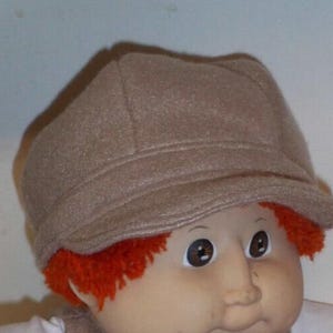 May include: A light brown newsboy cap worn by a doll with bright orange hair. The cap features a structured crown, a short curved brim, and a stitched band detail across the front. The doll has a round face with brown eyes and a light skin tone.