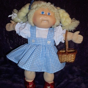 Może przedstawiać: Lalka Cabbage Patch Kid ubrana w niebiesko-białą sukienkę w kratę z białą bluzką i czerwonymi butami. Lalka ma blond włosy w warkoczach i trzyma brązowy wiklinowy koszyk.