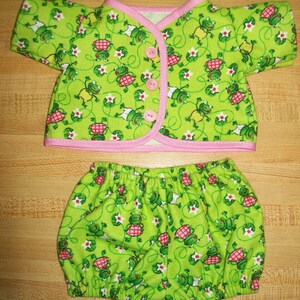 May include: A two-piece baby outfit featuring a lime green frog print. The top has short sleeves, pink trim, and pink buttons. The matching bloomers are gathered at the waist and legs. The fabric is covered in frogs, flowers, and vines.