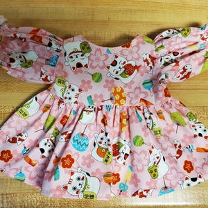Pode incluir: Um vestido rosa com mangas bufantes e uma borda de renda branca. O vestido é coberto por um padrão de gatos brancos, flores de cereja e caracteres japoneses.