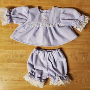 May include: A light purple baby doll outfit, comprising a top with lace trim on the sleeves and bodice, and matching bloomers with lace edging. The top has small flower-shaped buttons.
